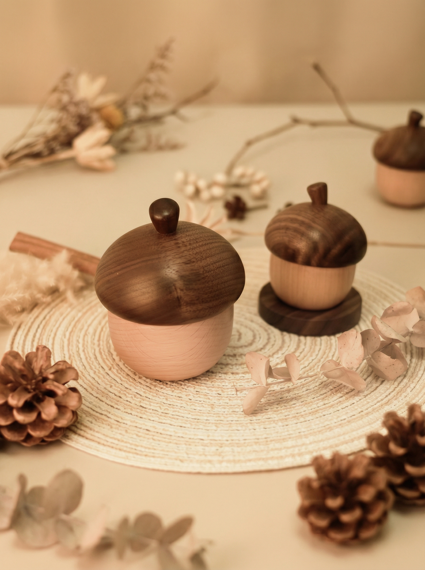 Håndlagde trebokser formet som eikenøtter med lokk, dekorert med kongler og tørkede blomster på flettet underlag.