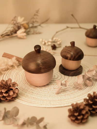 Håndlagde trebokser formet som eikenøtter med lokk, dekorert med kongler og tørkede blomster på flettet underlag.